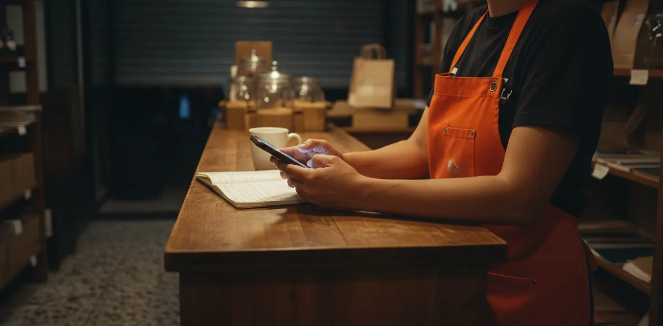 La dueña de un local, con delantal naranja, publica desde su teléfono en el mostrador después de cerrar